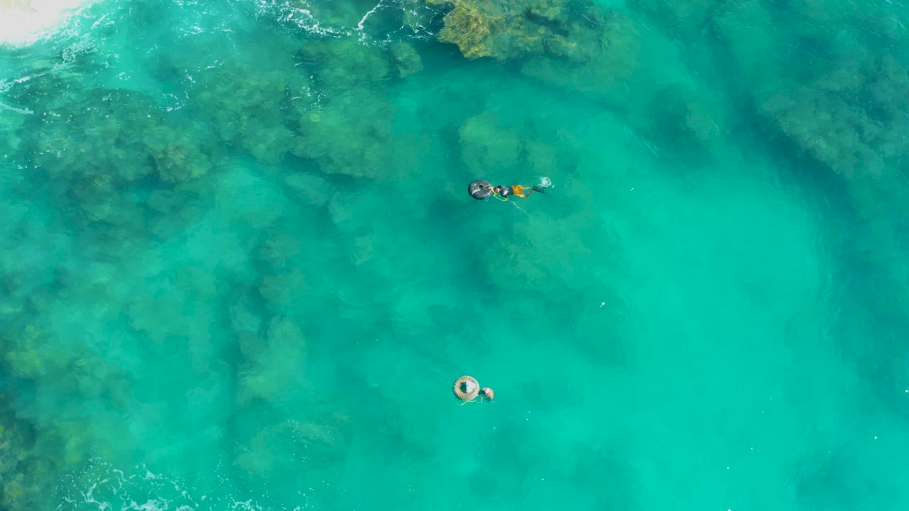 Overhead view in San Jose Del Cabo showing turquoise seawater, dark submerged reef patches near the top, and small swimmers separated across the frame.