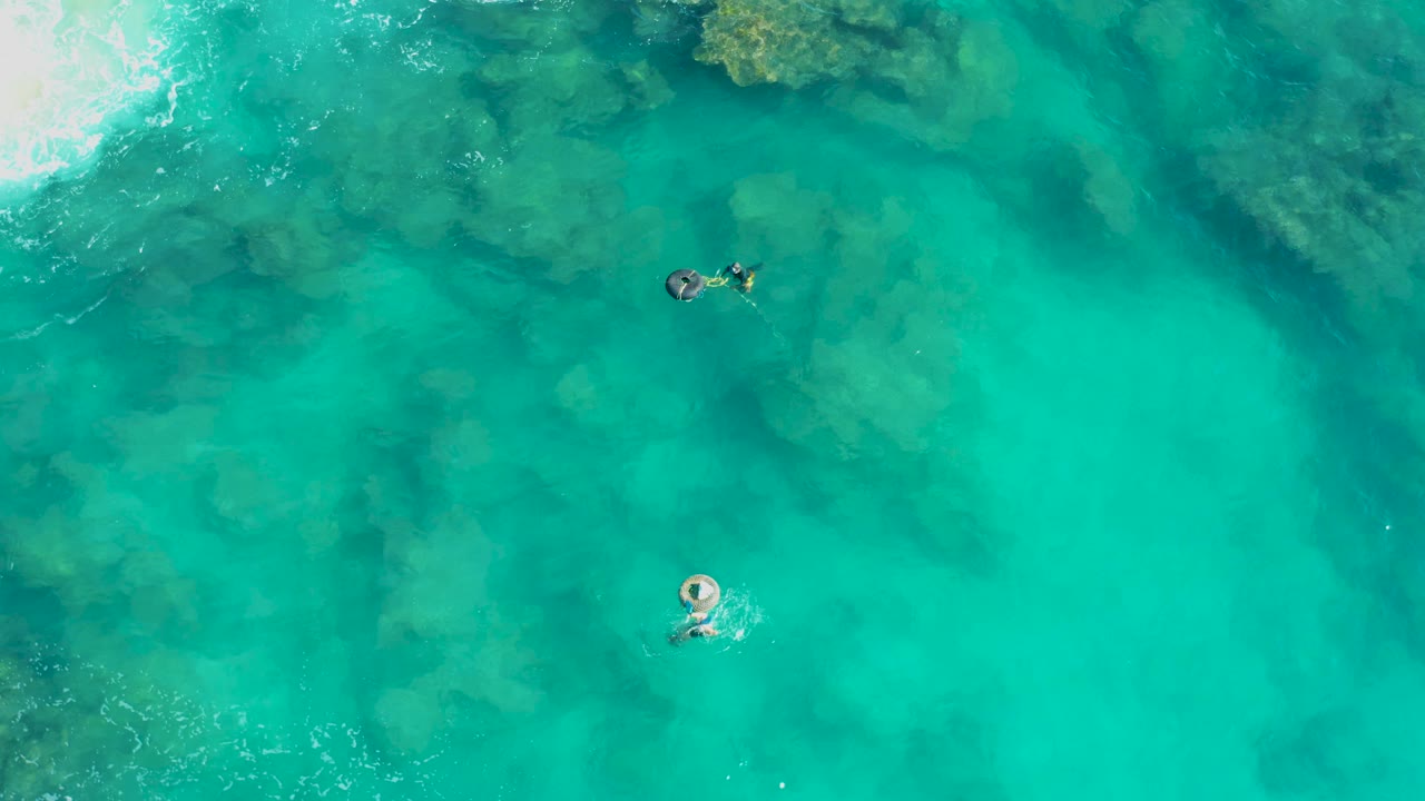 Overhead view in San Jose Del Cabo with two visible people farther apart in bright turquoise water, a pale ring lower in frame, a dark board near the upper swimmer, and mottled reef texture under the surface.