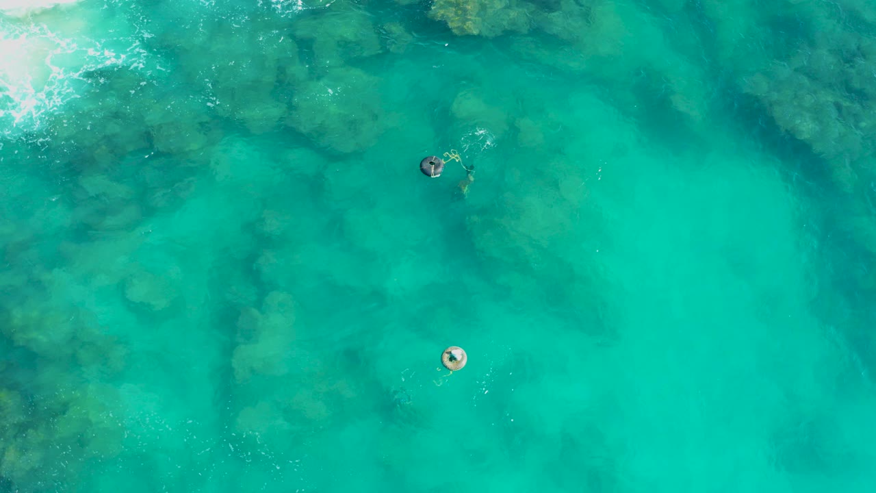 Overhead view in San Jose Del Cabo of two small swimmers separated in clear turquoise water, with submerged rock shapes and light foam near the upper left.