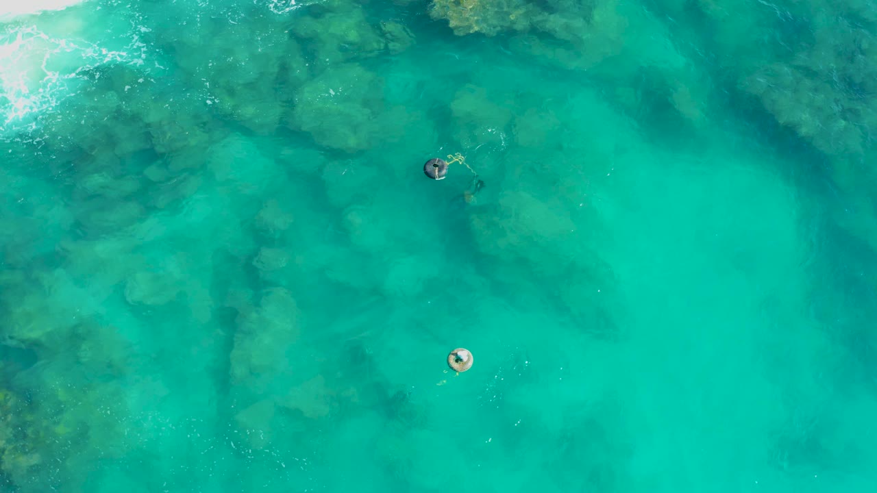 Top-down San Jose Del Cabo view with one swimmer near the upper middle and another lower in frame, both crossing clear turquoise water above uneven submerged rocks.