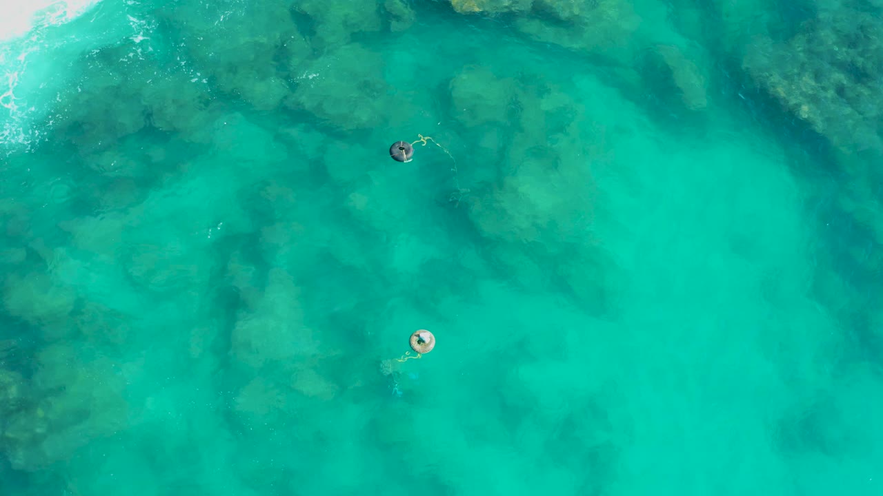 Aerial view in San Jose Del Cabo with a bright surf edge, two buoys over shallow turquoise water, underwater rock shapes, and no visible people.