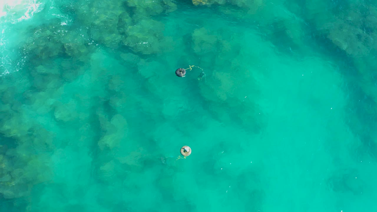 Overhead drone view in San Jose Del Cabo showing one person on a circular float and another swimmer separated in clear turquoise water with dark rocks below.