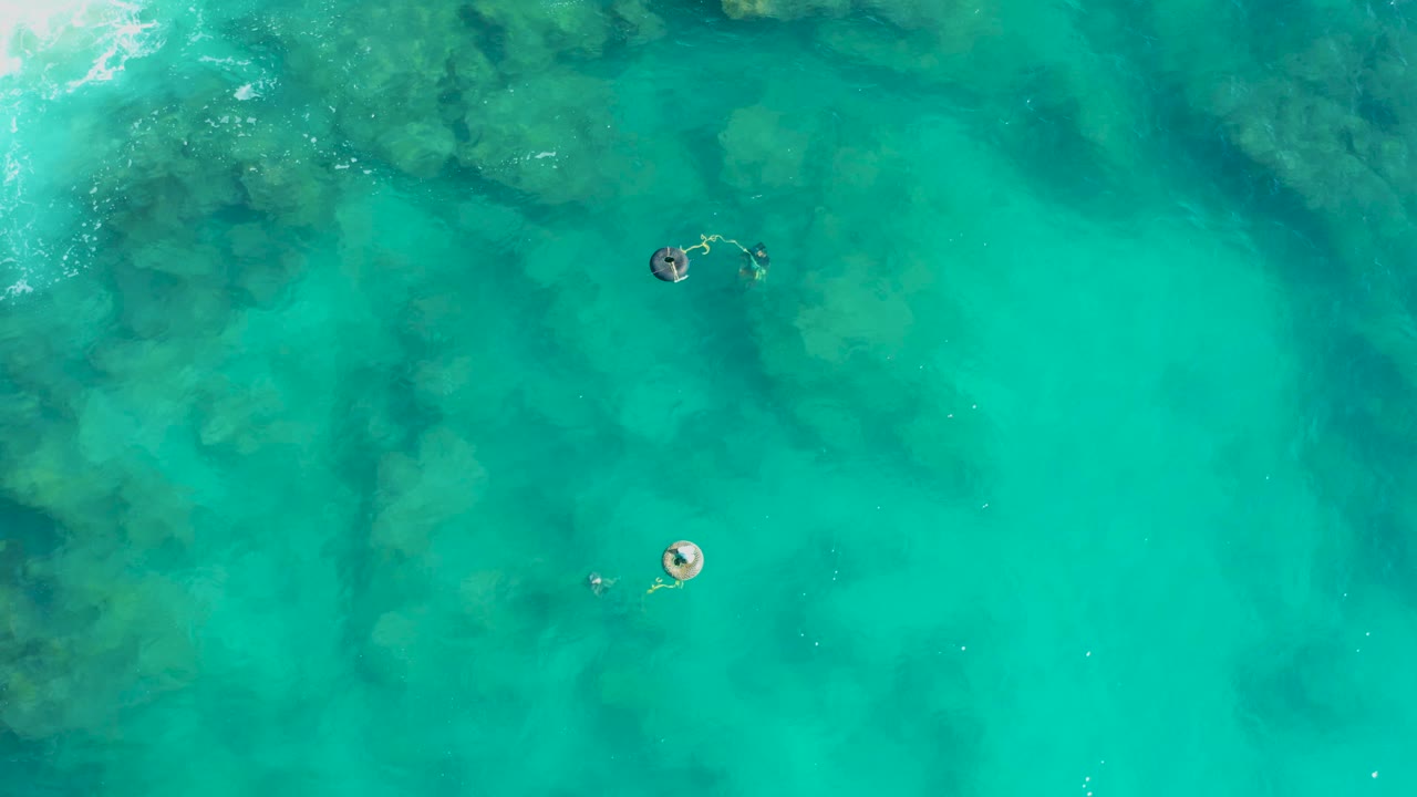 High overhead frame in San Jose Del Cabo showing a circular float to the lower right of a swimmer, with clear water shifting from aqua to darker rock patches.