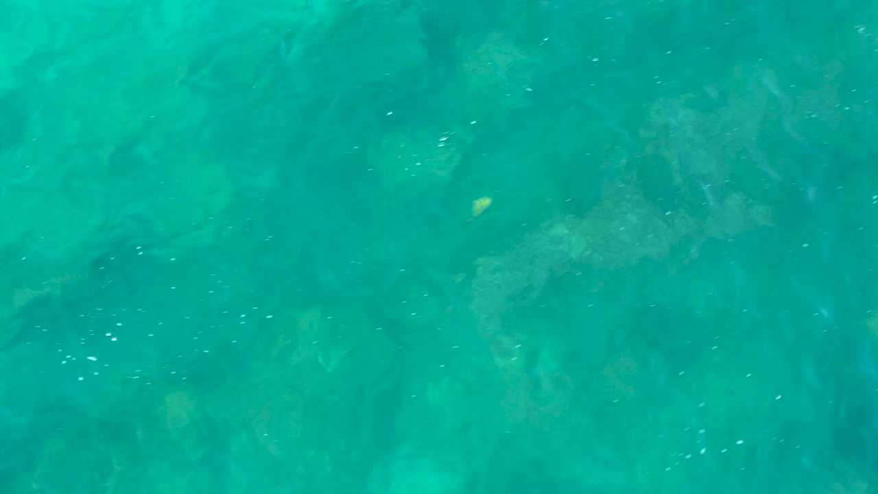 Turquoise water in San Jose Del Cabo exposes larger underwater rock forms and faint specks of bubbles, with no visible people.