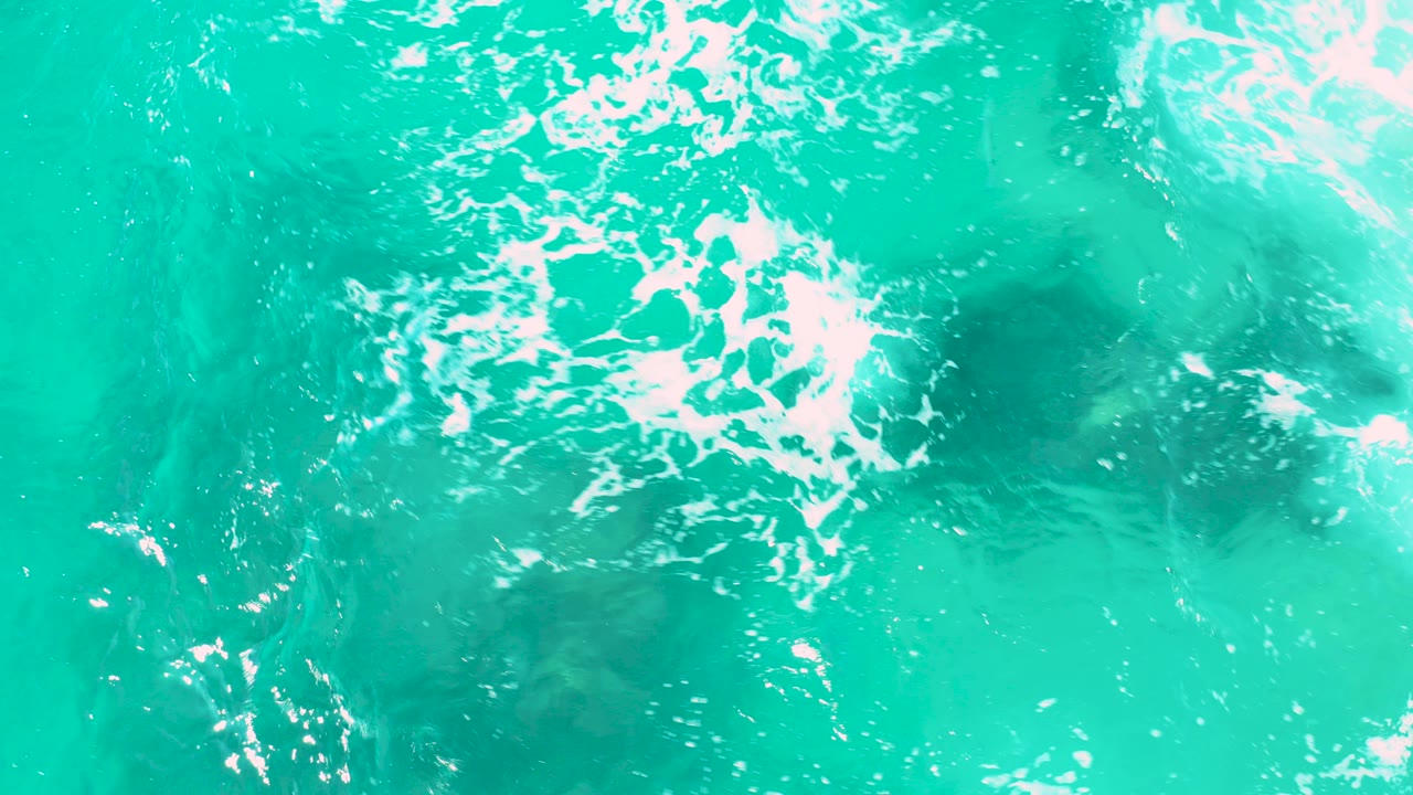 Top-down view of clear turquoise water in San Jose Del Cabo with white foam near the upper edge, darker seabed patches below, and no visible people.