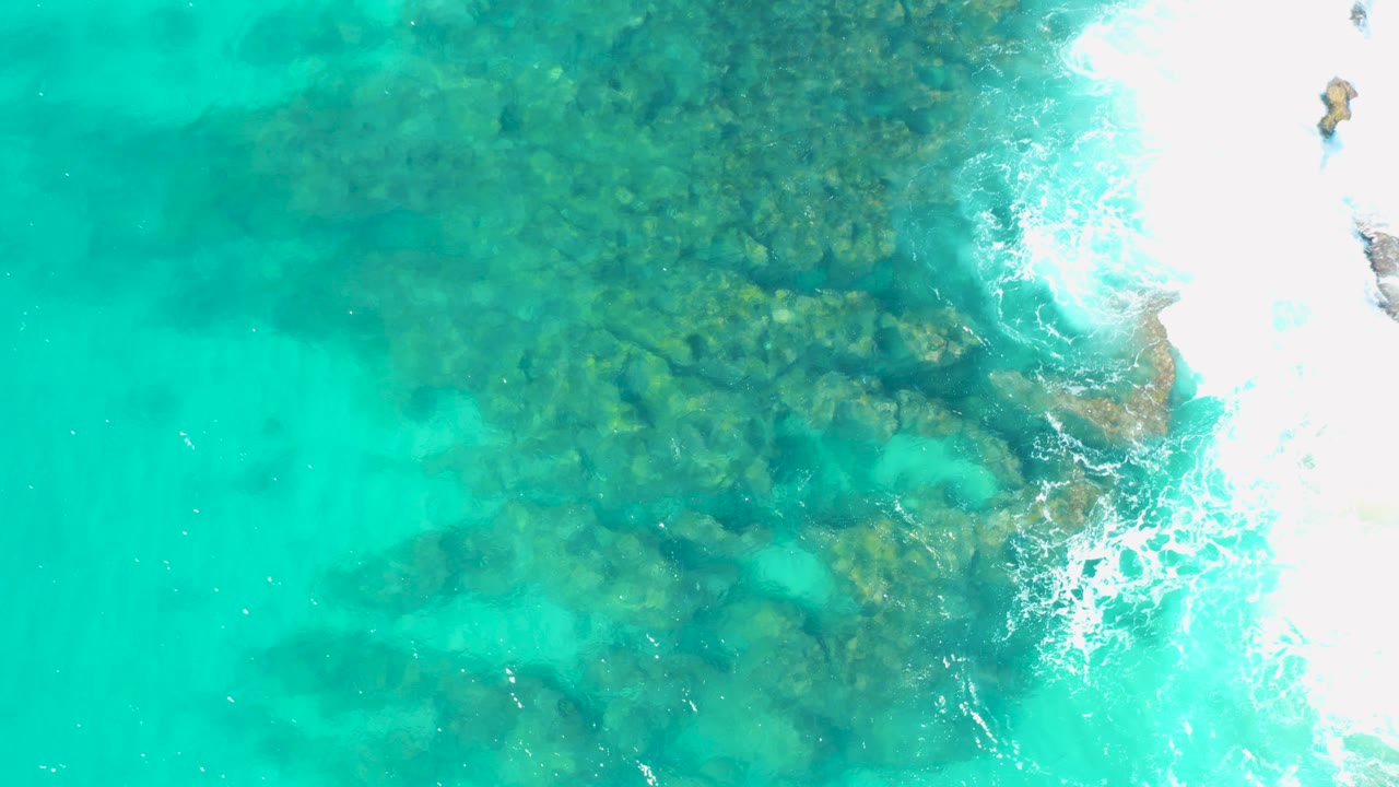 Overhead view in San Jose Del Cabo of turquoise water revealing rocky formations beneath the surface, with bright white surf entering from the right and no visible people.