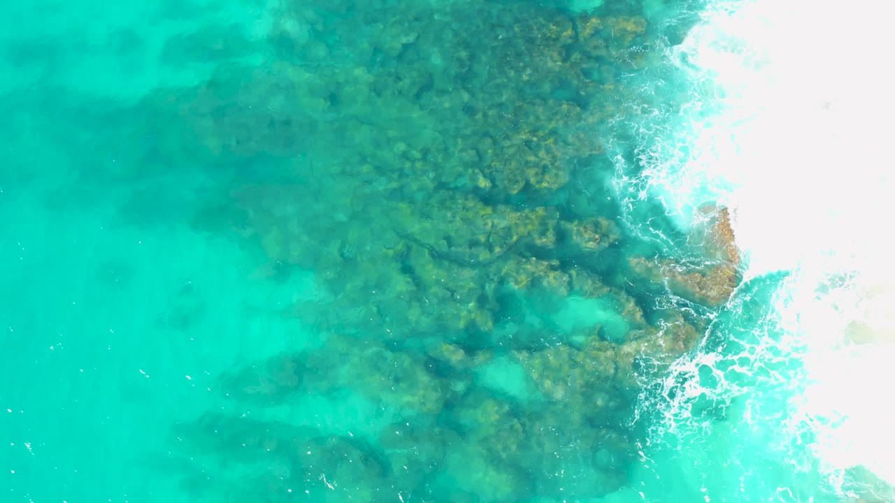 Overhead view in San Jose Del Cabo with reef-like rock texture under turquoise water and a larger burst of white surf along the right edge, with no visible people.