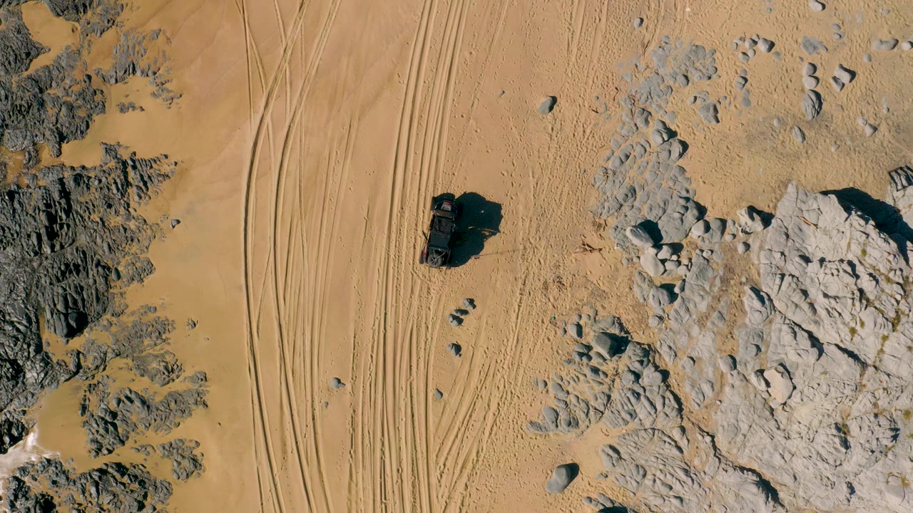 Overhead view of an off-road vehicle on sandy desert terrain beside rocks and tire tracks.