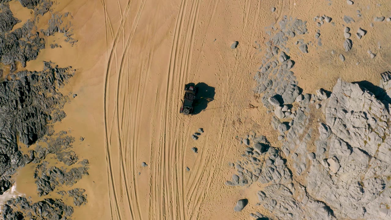 San Jose Del Cabo aerial frame with the off-road vehicle slightly farther along the sandy area, parallel tracks on one side, rock clusters on the other, and no visible people.