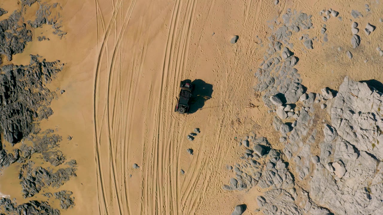 Drone-style overhead view in San Jose Del Cabo with the vehicle centered near compacted sand, narrow tire lines to the side, scattered stones, and no visible people.