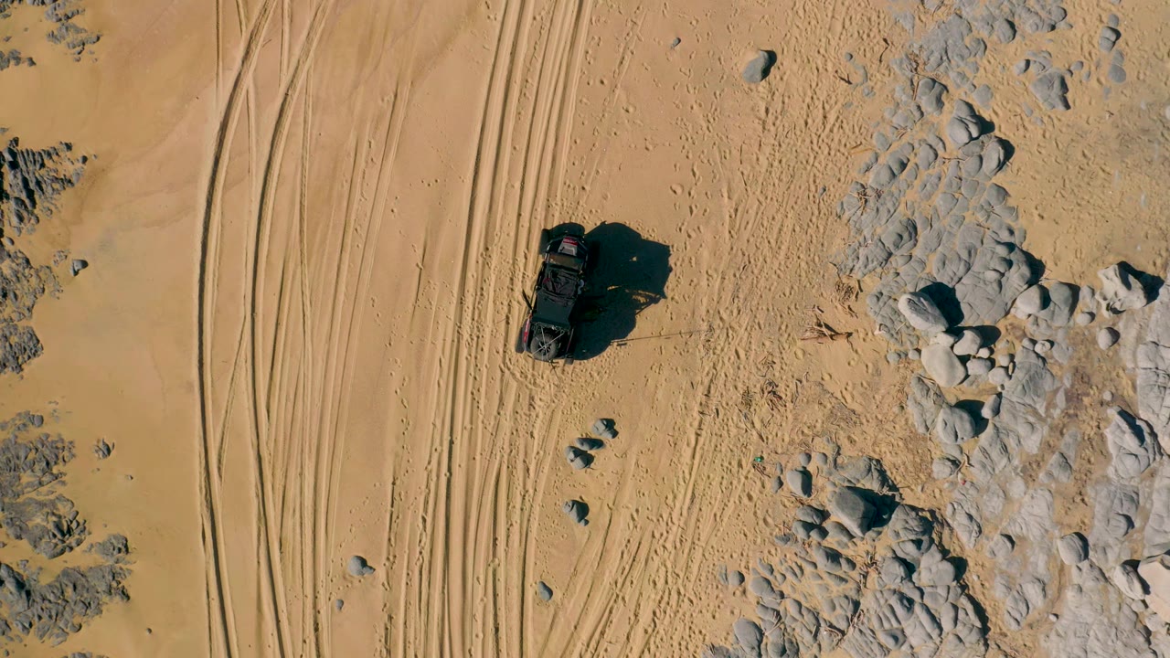 A top-down San Jose Del Cabo desert view shows the SUV slightly farther along the sandy path, with multiple tire lines to the left and pale rocks to the right, no visible people.