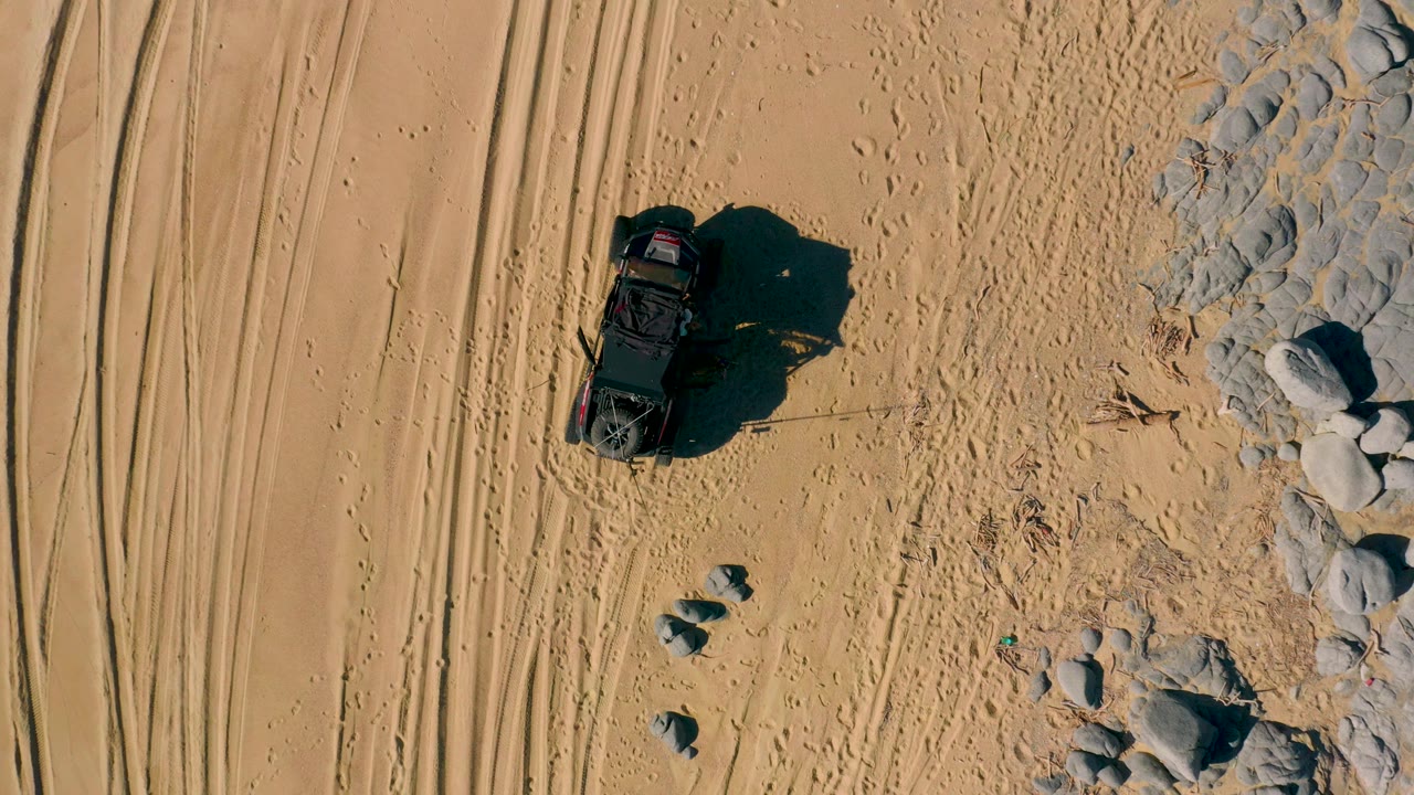 Aerial frame in San Jose Del Cabo, Baja California Sur, with the black off-road vehicle slightly lower in the sandy track area and pale rocks along the right edge, with no visible people.