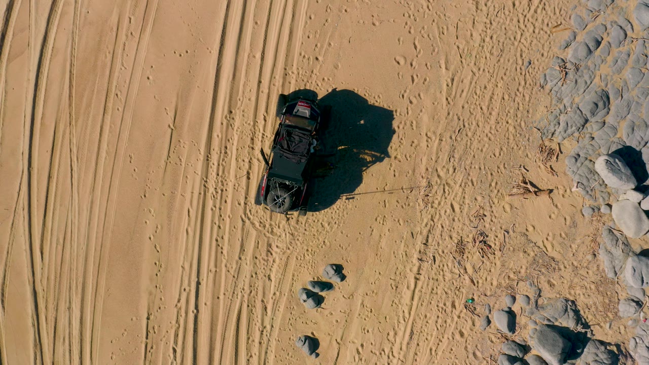 Top-down San Jose Del Cabo desert view of the black off-road vehicle moving through marked sand with tire lines to the left and scattered rocks to the right, with no visible people.