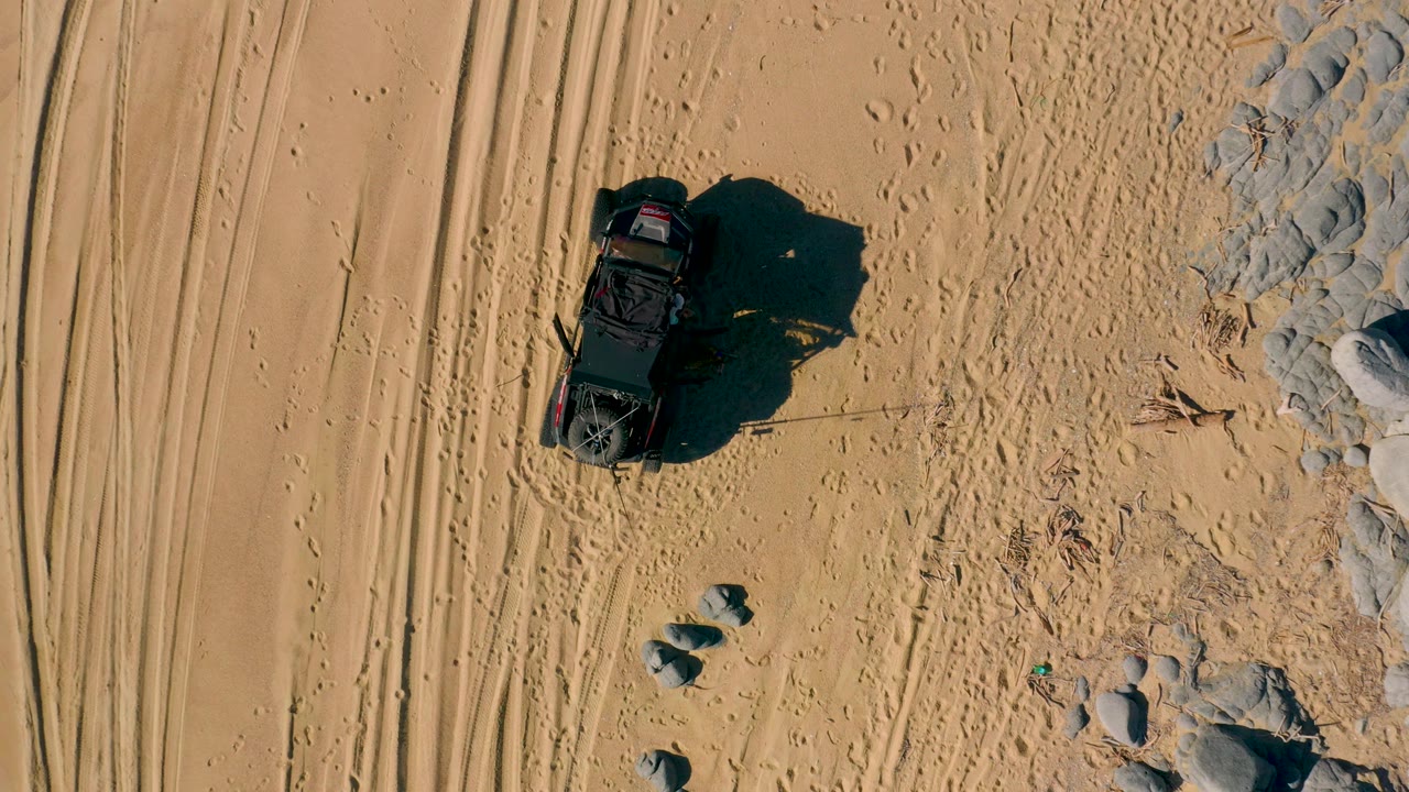 Overhead shot in San Jose Del Cabo, Baja California Sur, showing the black off-road vehicle closer to the center-left of frame, with deep shadow, tire tracks, footprints, and rocks, with no visible people.