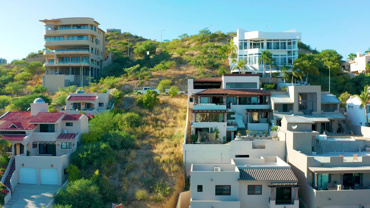 San Jose Del Cabo hillside residences fill the frame in bright daylight, with white and red-roofed homes, dry green brush, and no visible people.