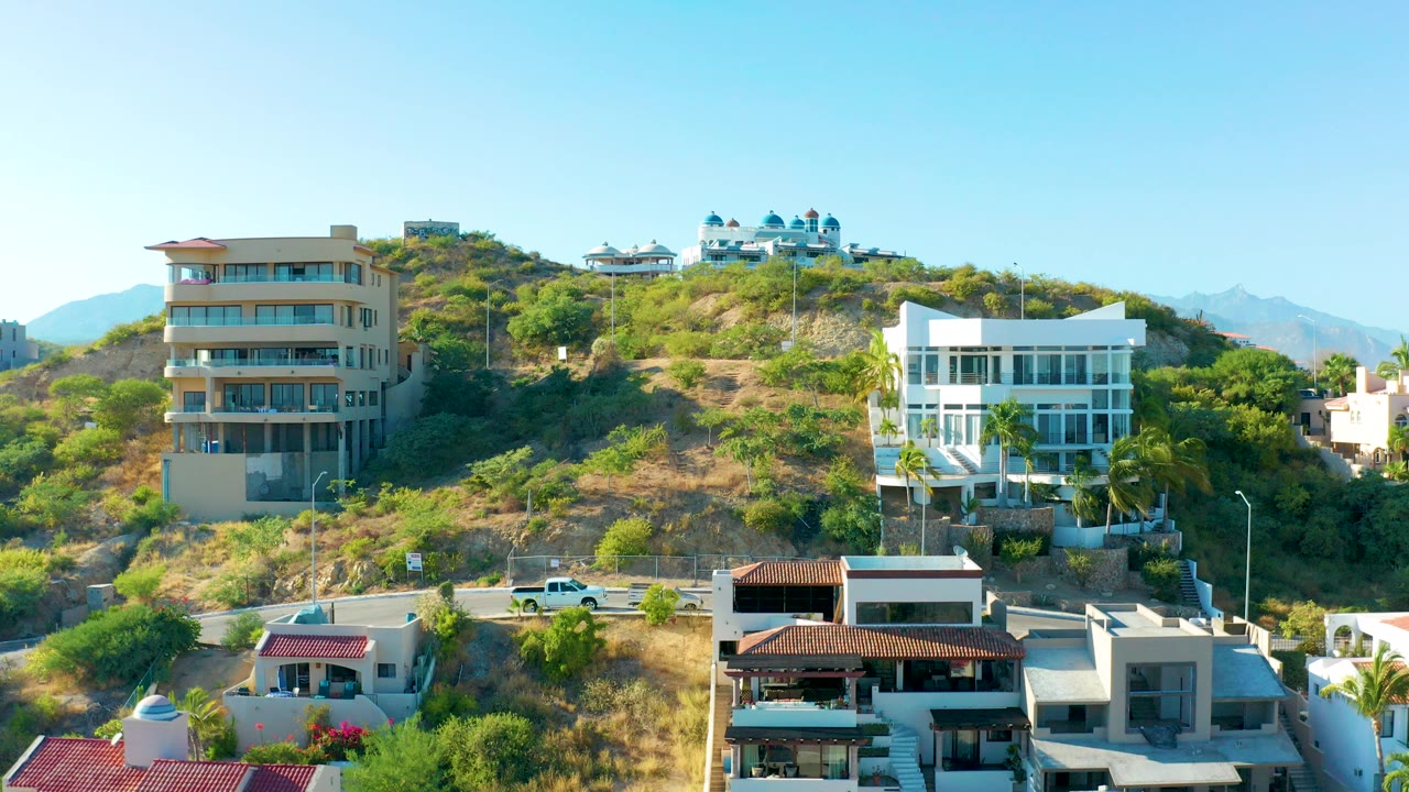 San Jose Del Cabo hillside residential area with tan and white modern buildings, a road at the base, dry vegetation, distant mountains, clear blue sky, and no visible people.