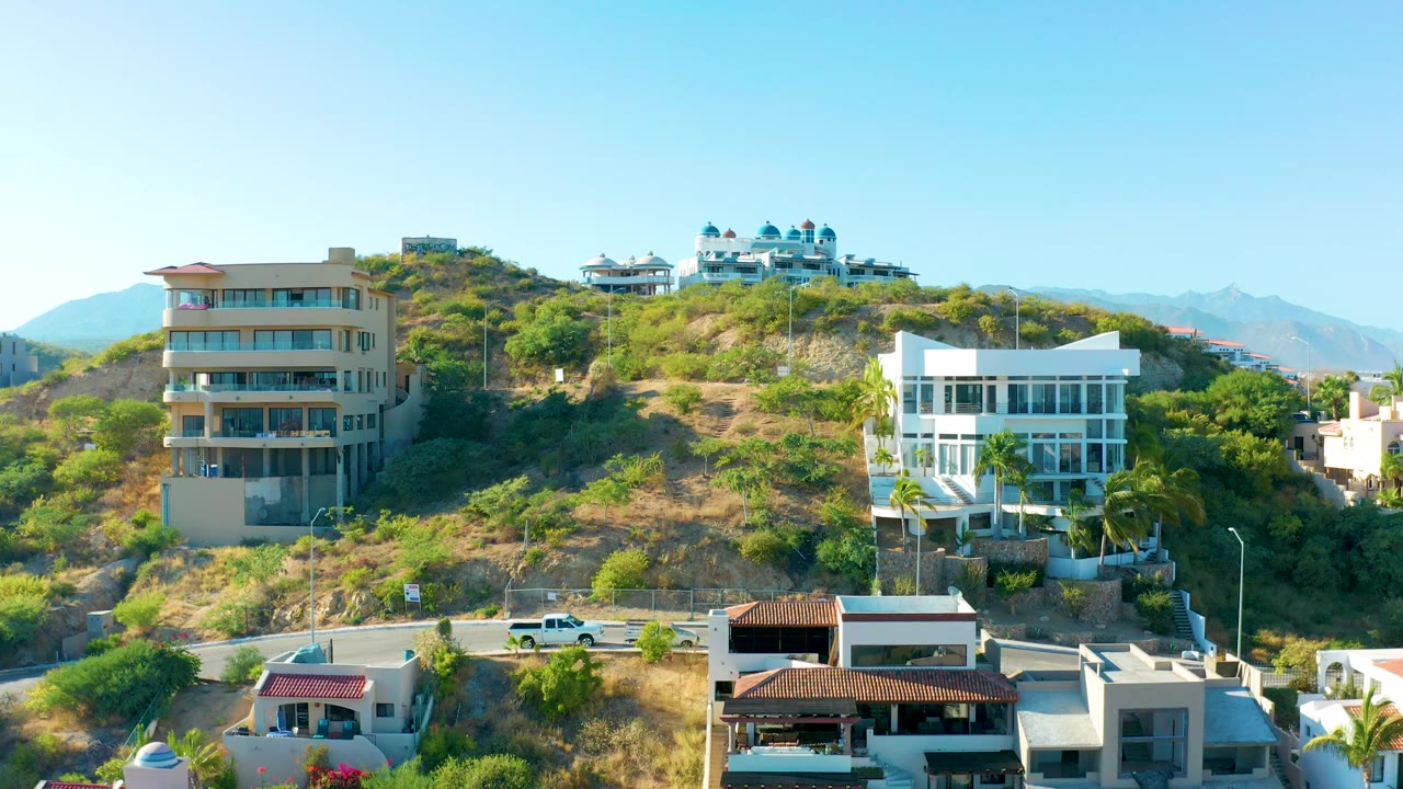San Jose Del Cabo hillside neighborhood showing a tan building on the left, a white house on the right, a road crossing the lower slope, dry brush, and no visible people.