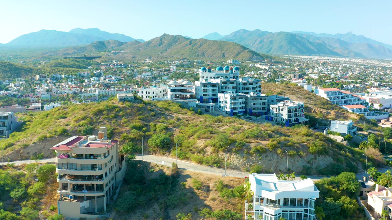 Wide aerial frame of San Jose Del Cabo showing hillside residences above a winding road, a cityscape across the valley, distant mountains, clear sky, and no visible people.