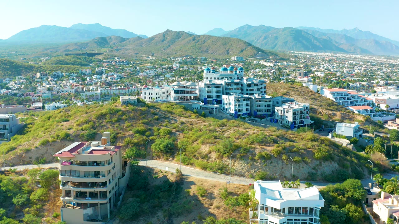 High aerial frame of San Jose Del Cabo with a curved hillside road below, clustered residential buildings on the slope, a broad urban area, blue mountain ridges, and no visible people.