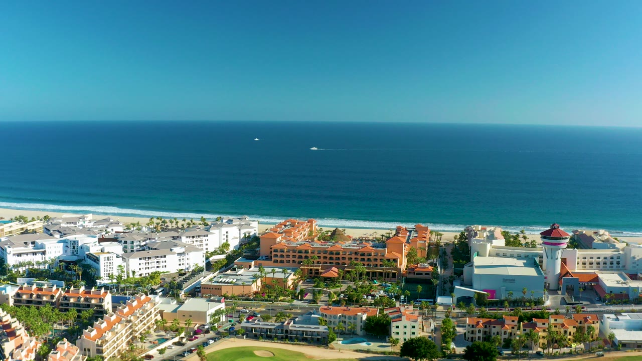 Opening aerial frame over San Jose del Cabo with white and orange-roofed buildings below a sandy beach, blue ocean, clear sky, and no visible people.