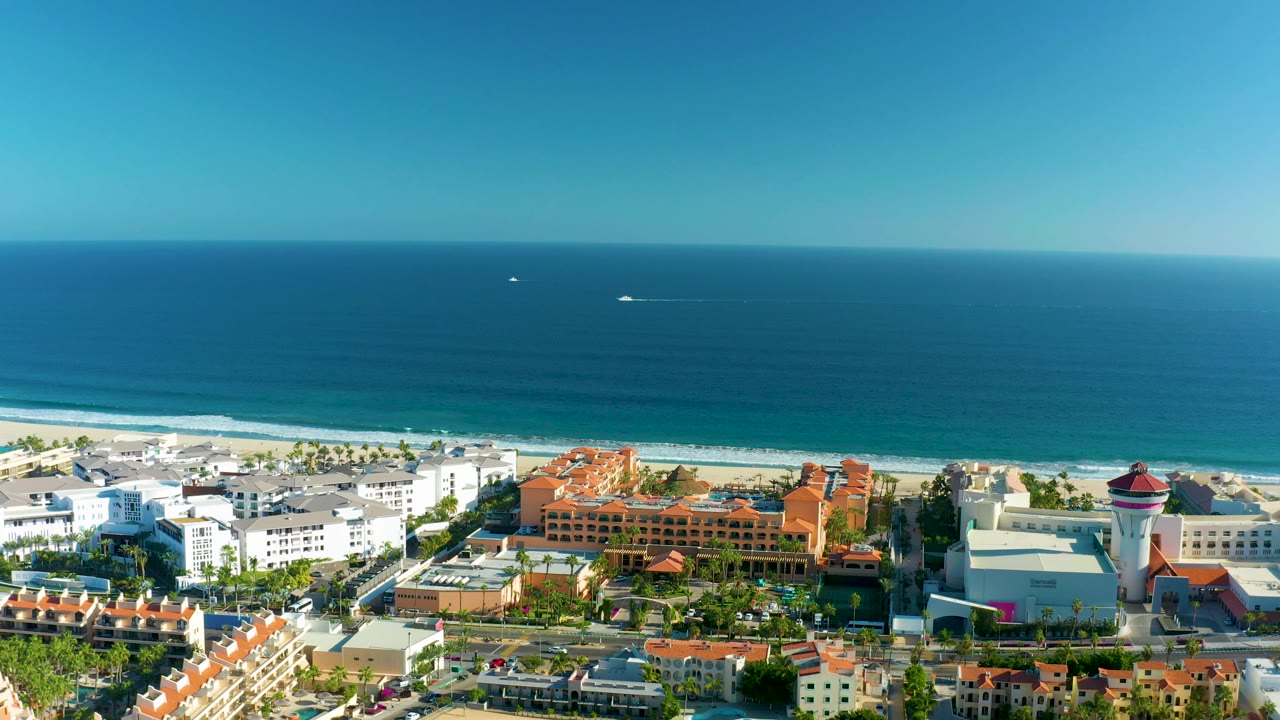 Mid-clip aerial view over San Jose del Cabo with the camera farther along the shoreline, white resort-style buildings in the lower frame, a long beach band, calm blue ocean, and no visible people.