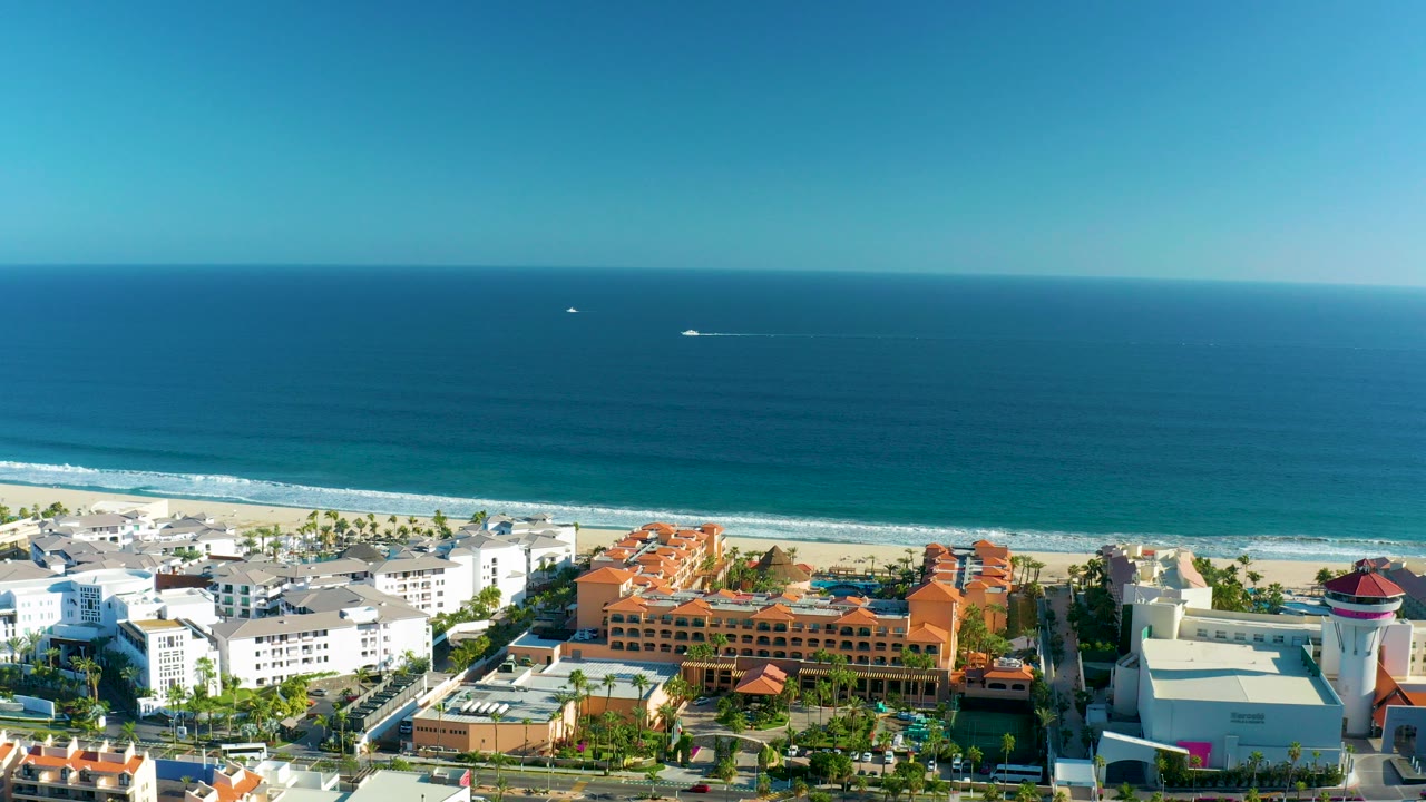 Wide aerial frame over San Jose Del Cabo showing blue ocean, a sandy beach, orange-roofed and white coastal buildings, palm trees, and no visible people.
