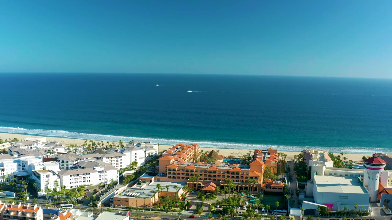 Aerial view in San Jose Del Cabo with the ocean horizon high in frame, small boats offshore, a pale sandy beach, and waterfront buildings below with no visible people.