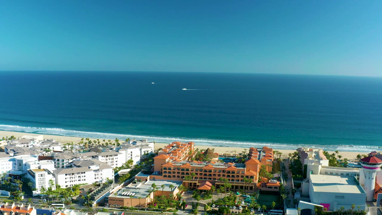 High aerial frame over San Jose Del Cabo showing a broad band of ocean, breaking waves at the sandy shore, white beachfront structures, orange roofs, and no visible people.