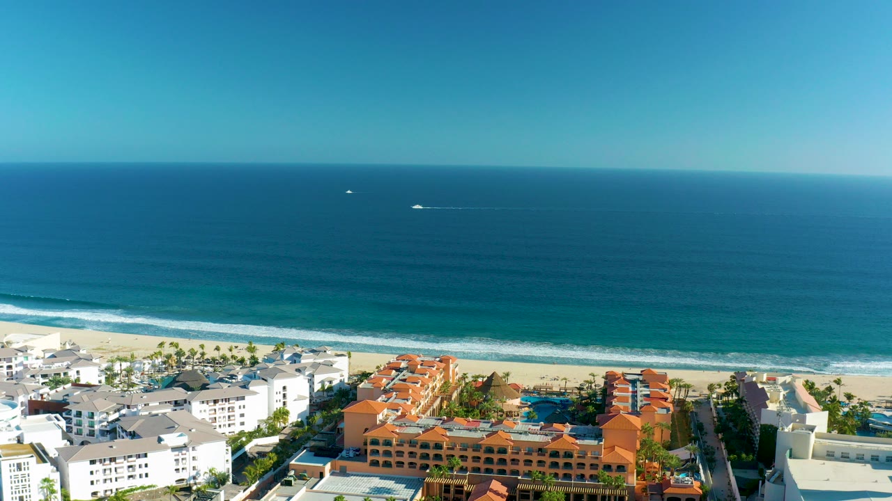 Aerial frame over San Jose Del Cabo showing red-orange and white beachfront buildings below a sandy beach, breaking surf, blue ocean, clear sky, and no visible people.