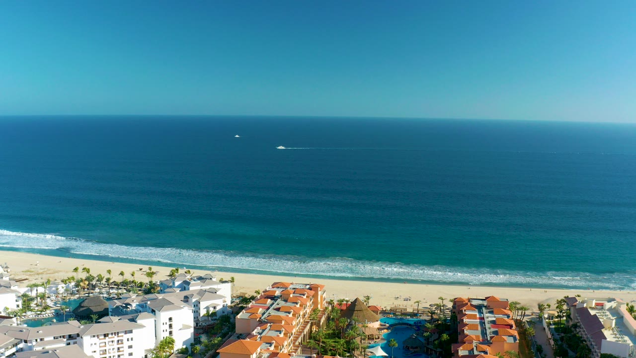 At 0:00, an aerial view over San Jose Del Cabo shows blue ocean, a sandy resort beach, palm trees, white surf, and a small distant boat with no visible people.