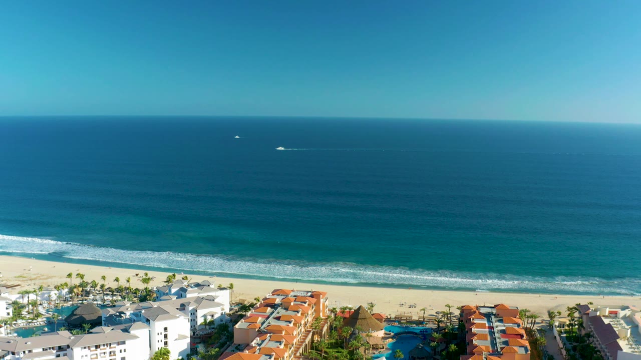 At 0:02, a high aerial frame shows the boat and its white wake on the ocean, with turquoise water near shore, a sandy beach, resort roofs, and no visible people.