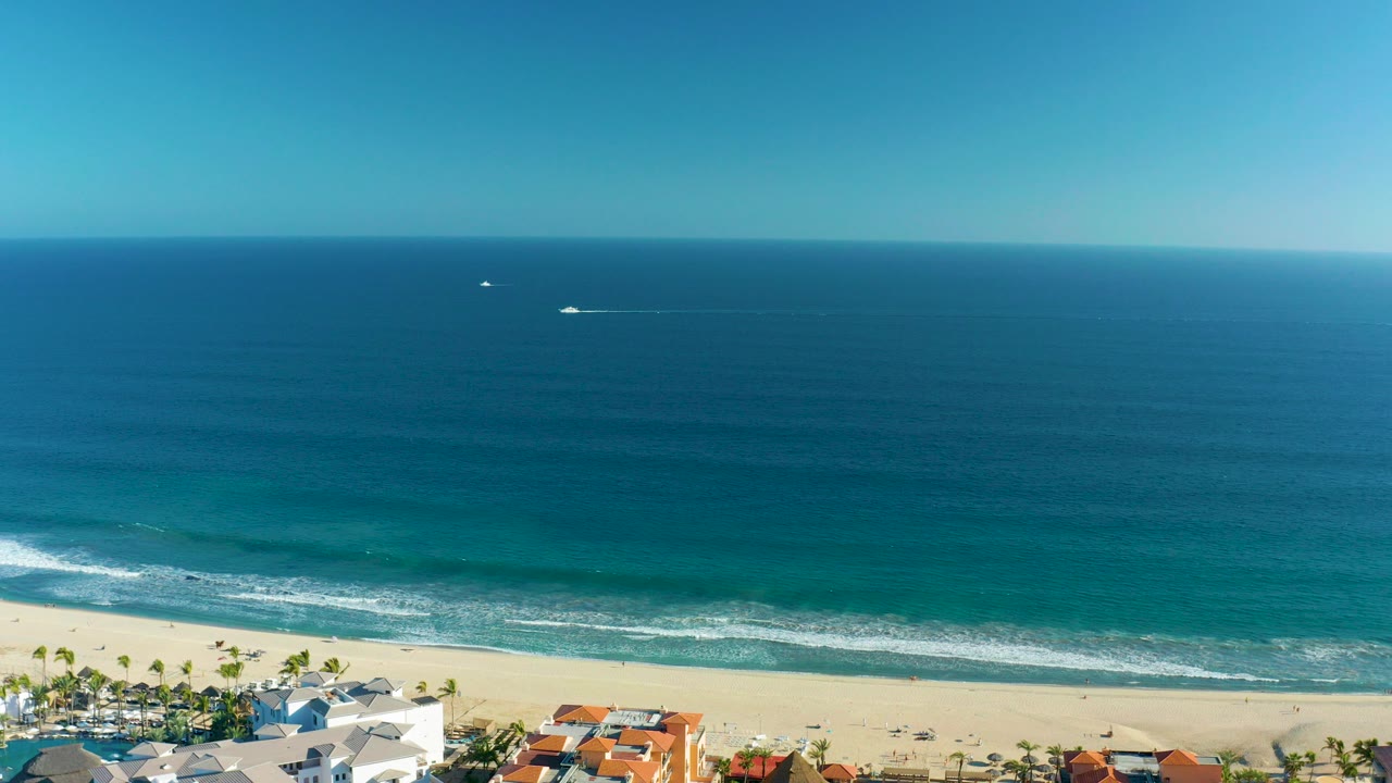 Opening aerial view of San Jose del Cabo coastline with deep blue ocean, turquoise shallows, white surf lines, sandy beach, palms, coastal homes, small boats offshore, and no visible people.