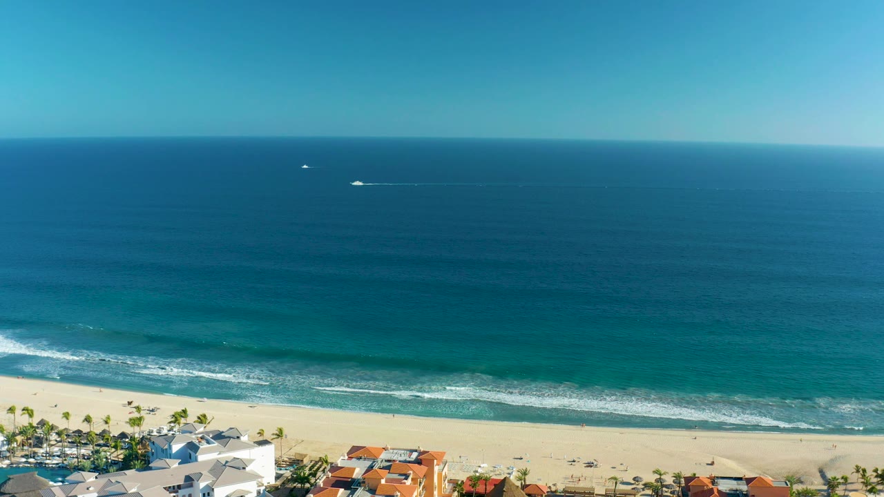 San Jose del Cabo aerial shoreline with turquoise water near the sand, a long white wave breaking parallel to the beach, palm-lined homes below, distant boats, and no visible people.