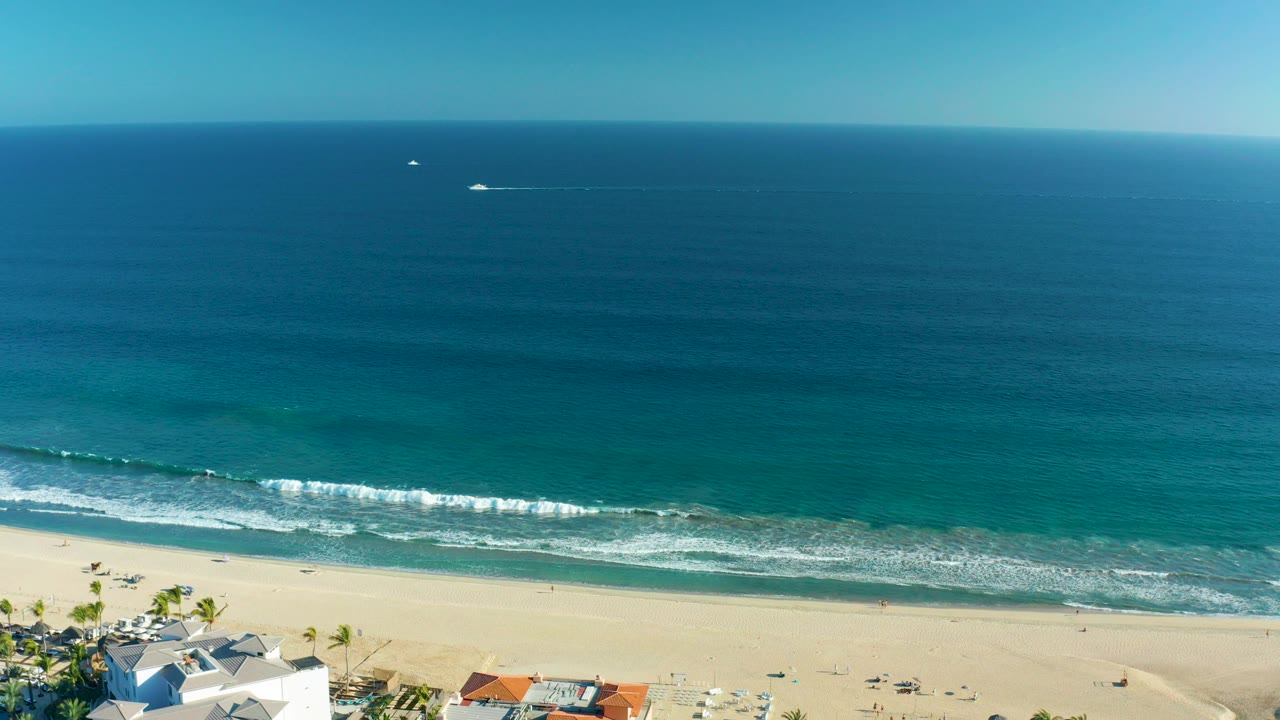 High aerial frame over San Jose Del Cabo with deep blue ocean, turquoise shallows, white surf, sandy beach, beachfront rooftops, palms, and tiny beachgoers visible near the lower edge.