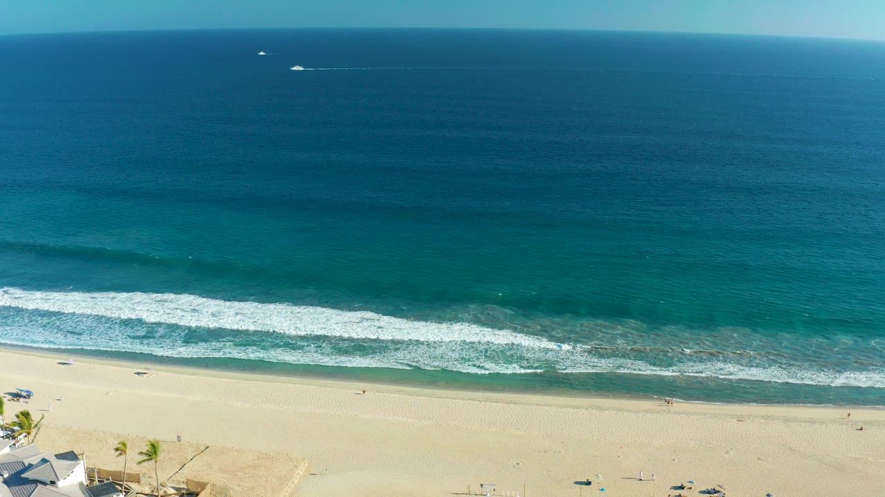 Aerial frame over San Jose Del Cabo showing blue ocean, a white surf line, pale sand, small beachfront structures at lower left, and tiny people on the beach.