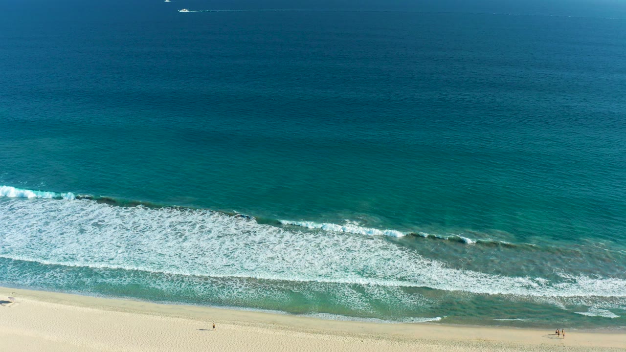 Aerial view over San Jose del Cabo shoreline with turquoise water, white surf, pale sand, tiny beachgoers near the lower edge, and small boats near the horizon.