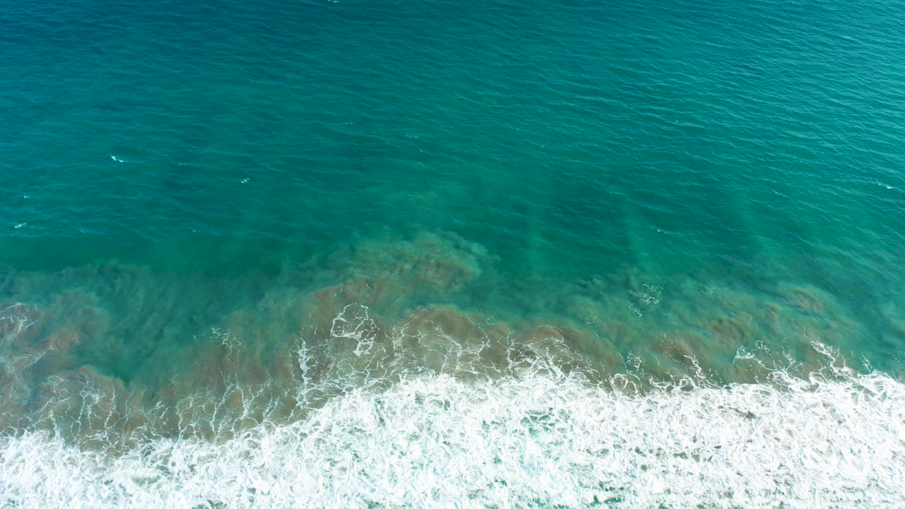 Top-down turquoise ocean with white surf over a shallow seabed.