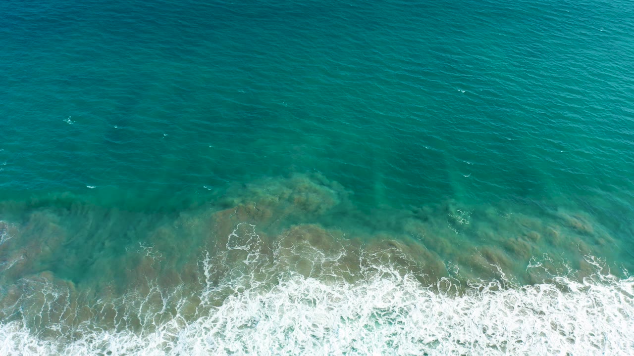 Top-down San Jose Del Cabo ocean frame with turquoise water, a dense white surf band, and darker underwater rock shapes visible beneath, with no visible people.