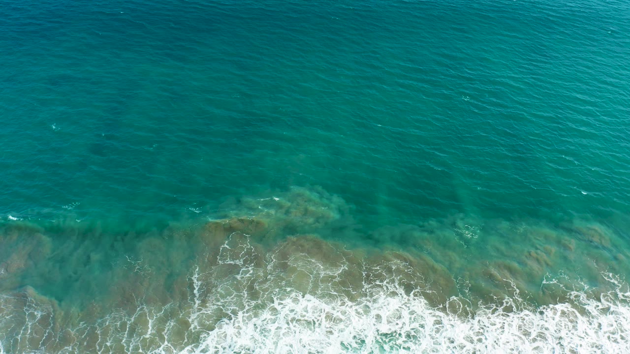 Aerial San Jose Del Cabo view with mostly open turquoise water and a receding white wave line near the bottom, with no visible people or vessels.