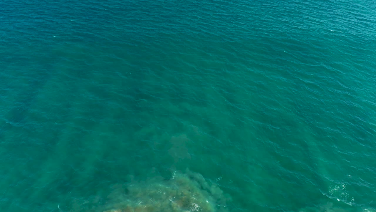 Clear blue-green ocean surface fills the frame in San Jose del Cabo, with faint underwater shapes and small wave texture, no visible people.