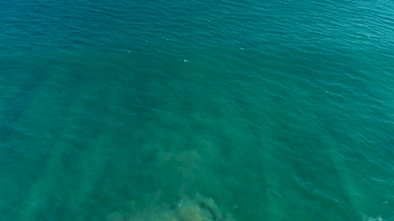 Turquoise ocean water in San Jose del Cabo shows a pale submerged rock area near the lower center beneath wind-made surface ripples, with no visible people.