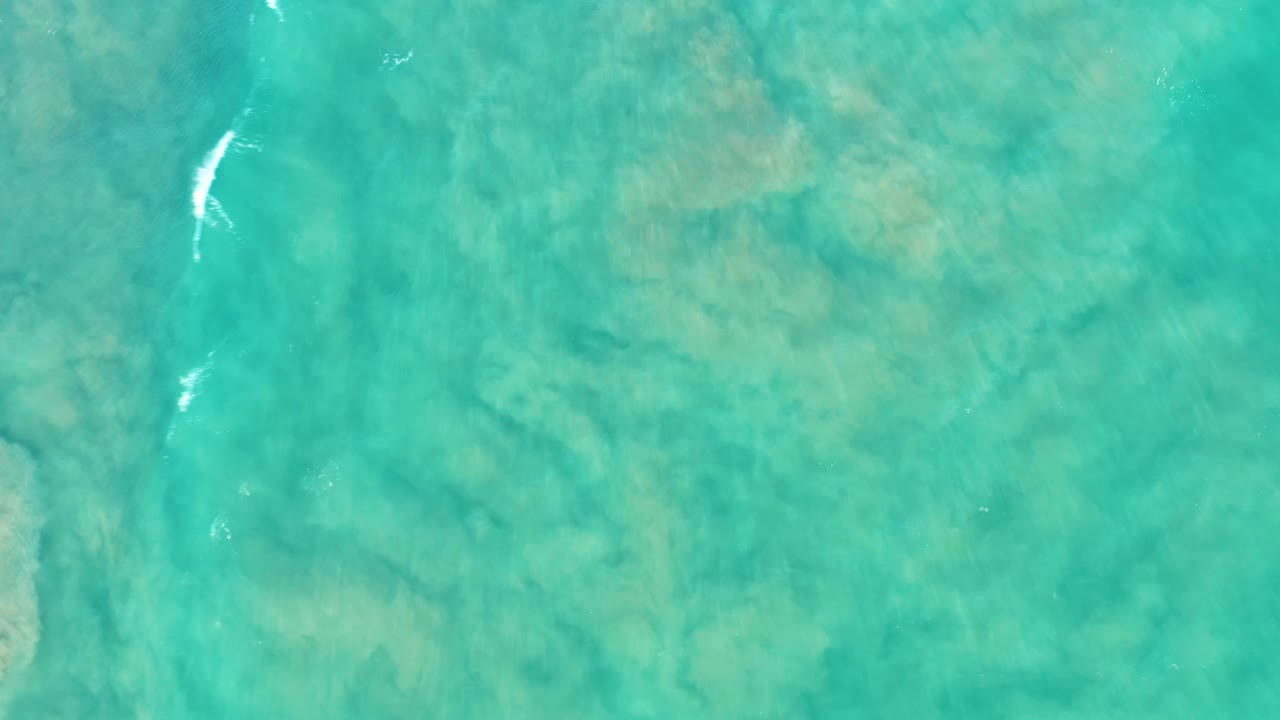 Clear green-blue ocean surface in San Jose Del Cabo with sand patterns under the water, a thin white foam mark near the upper left, and no visible people.