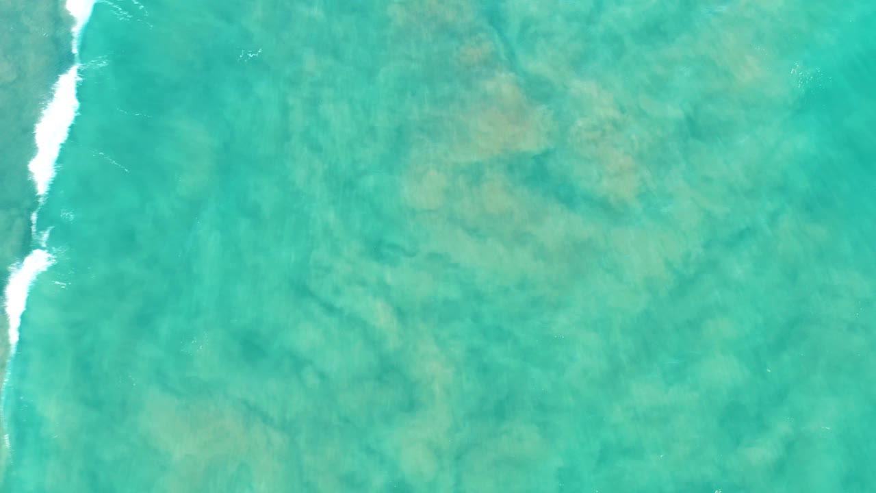 Top-down San Jose Del Cabo water view with a brighter white foam streak near the upper right, mottled sand below, and no visible people.