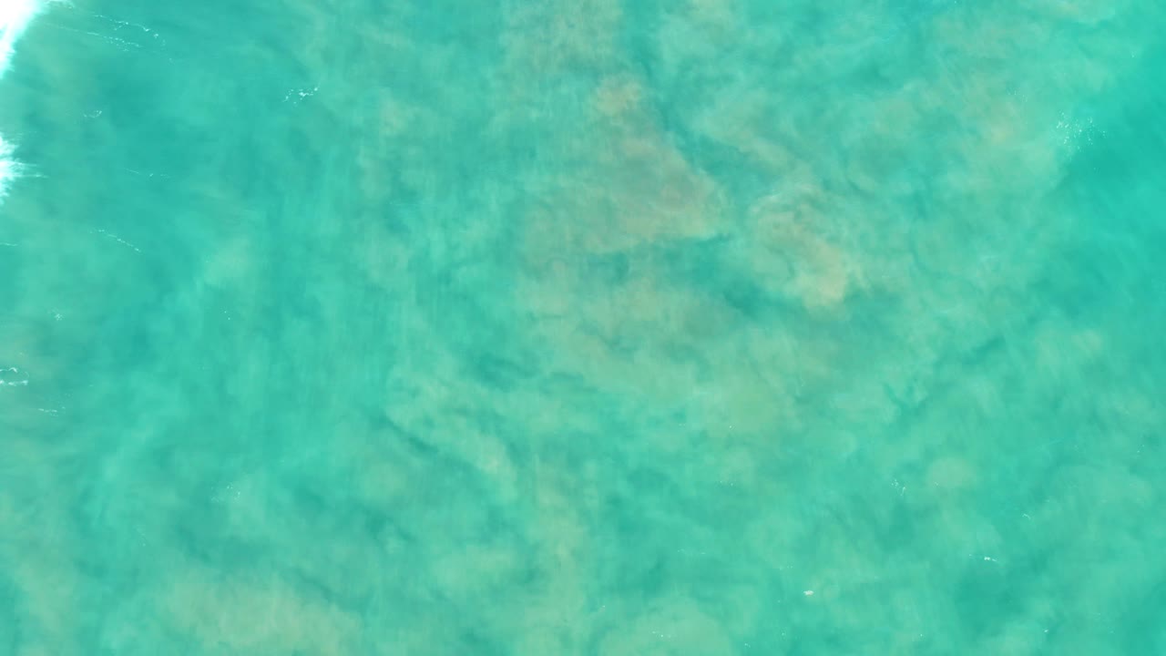 Aerial view over San Jose Del Cabo turquoise water with faint foam touching the left edge, soft sandy patches below, and no visible people.