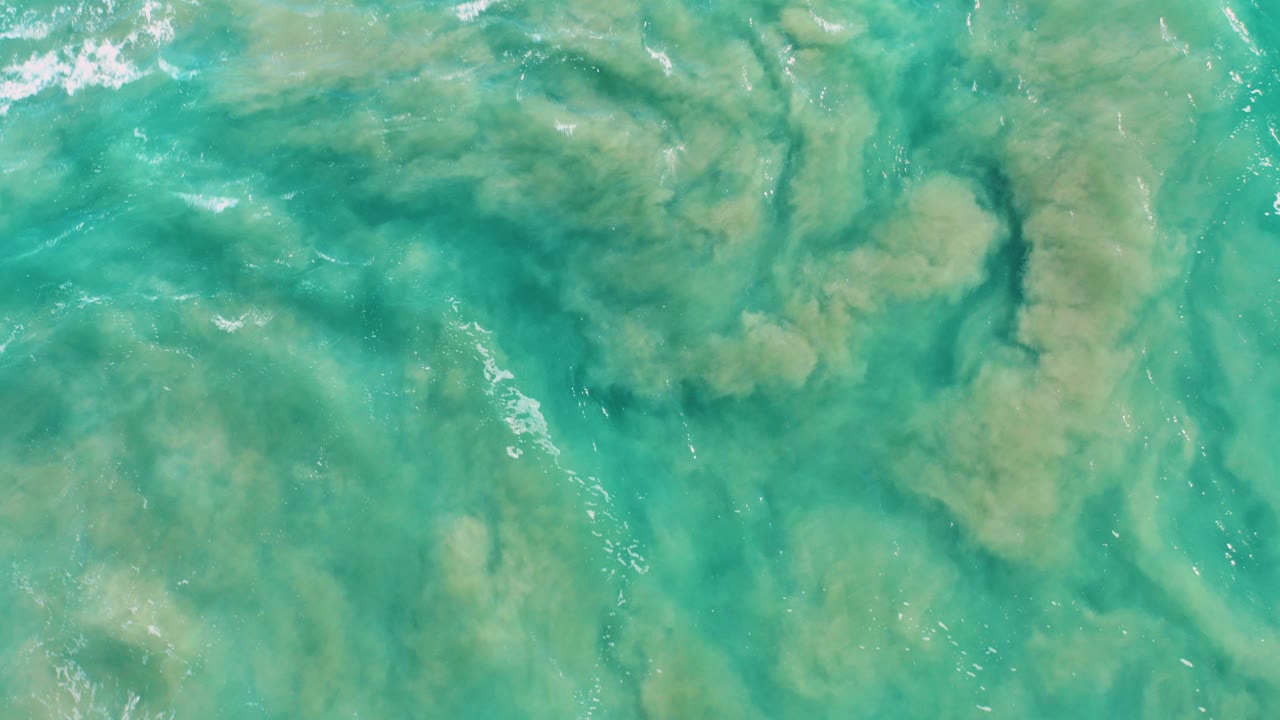 Top-down view of turquoise ocean water in San Jose del Cabo with green algae or sediment patches, faint ripples, and no visible people.