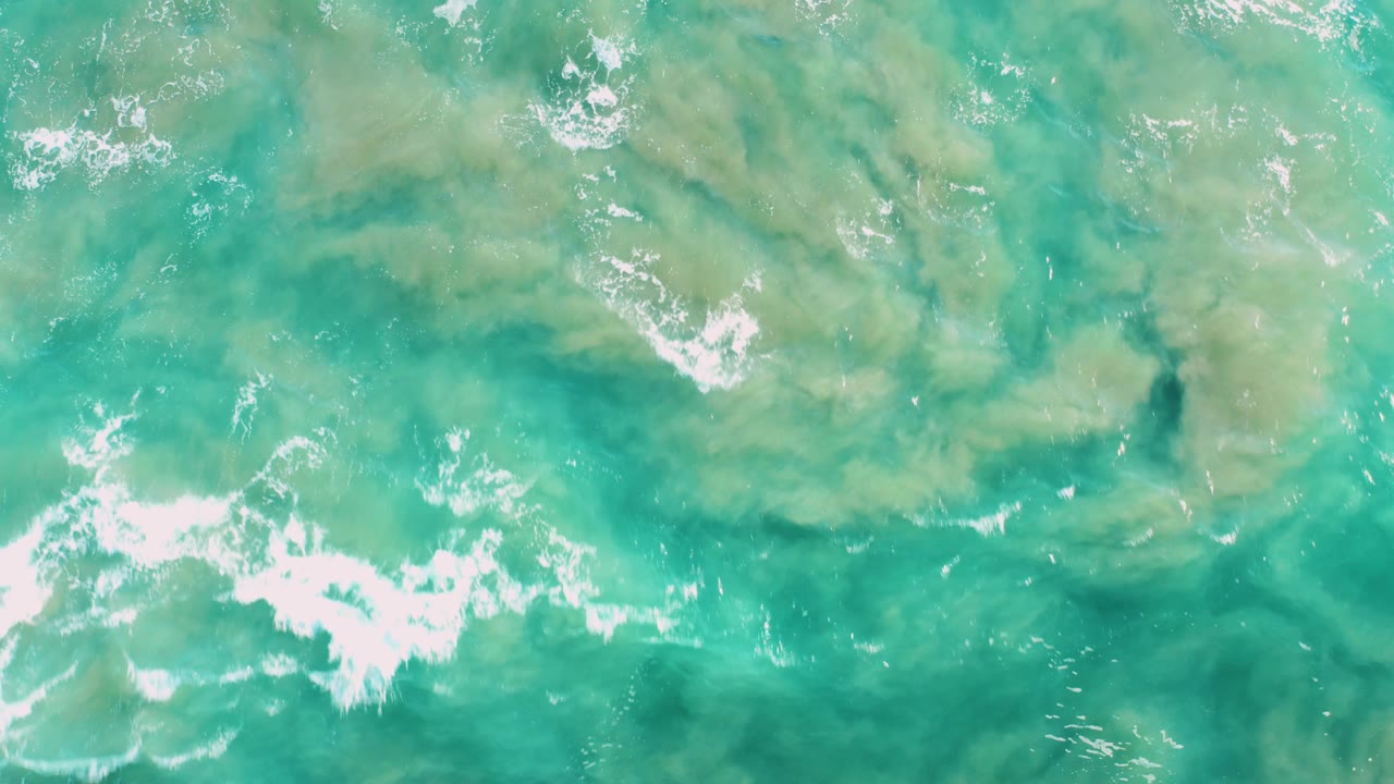 Ocean surface with turquoise water, pale green patches, and foam at the lower left.
