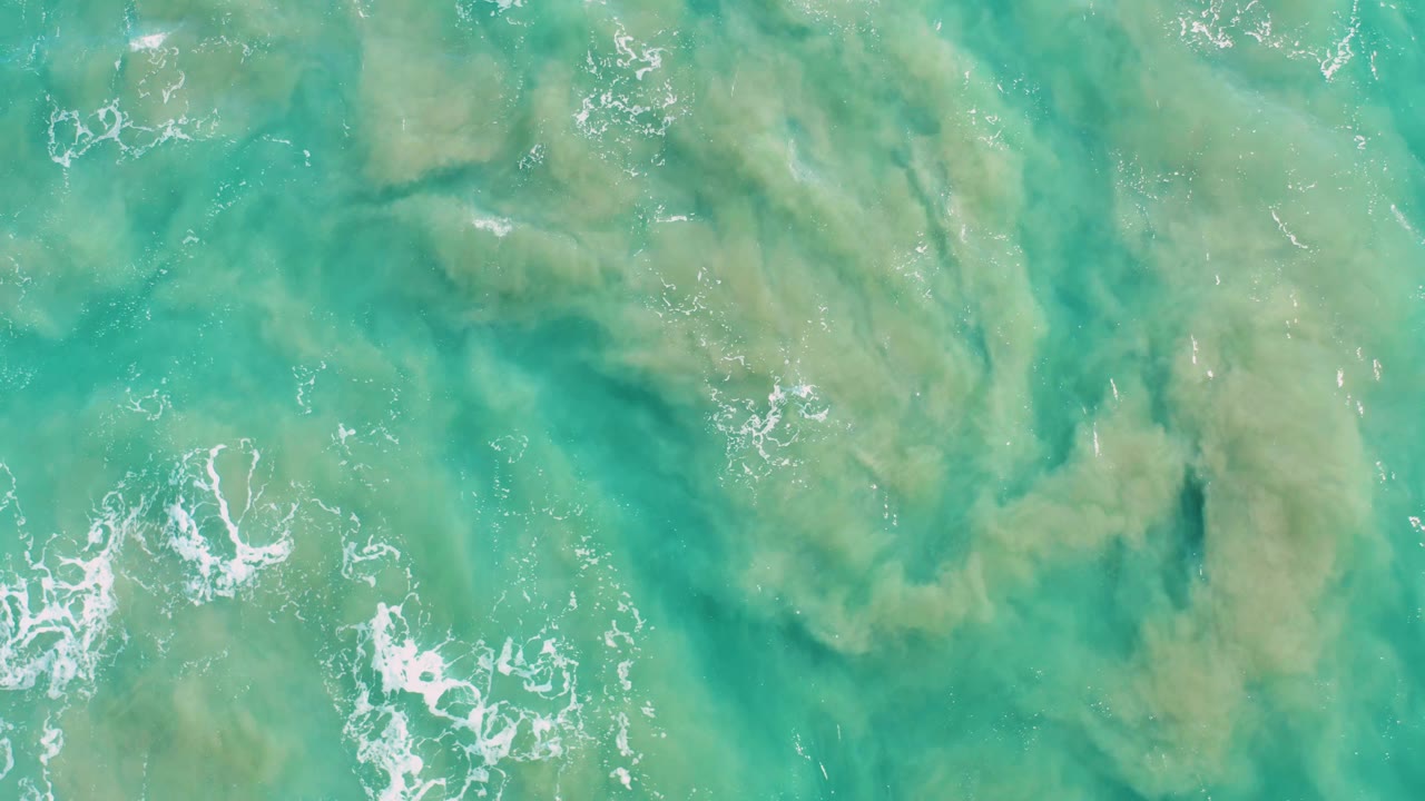 Aerial ocean surface in San Jose Del Cabo showing a broad greenish-brown sediment cloud under turquoise water, small foam streaks, and no visible people.