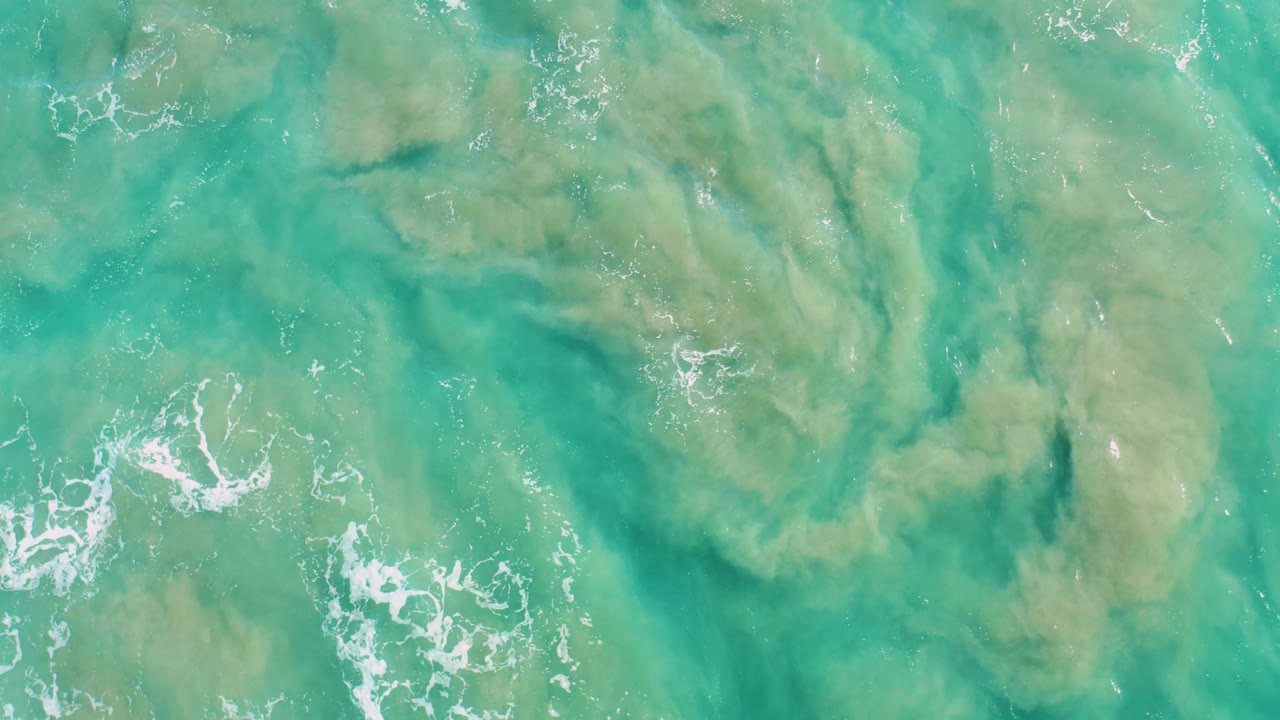 Straight-down San Jose Del Cabo ocean frame with white foam flecks over turquoise water and muted green-brown algae or sediment visible below, with no visible people.