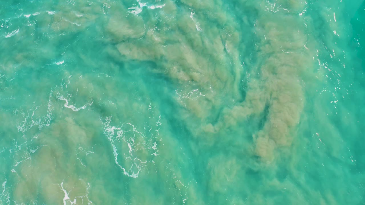 Clear green-blue ocean surface over pale sandy seabed in San Jose del Cabo, with foam flecks drifting near the center and no visible people.