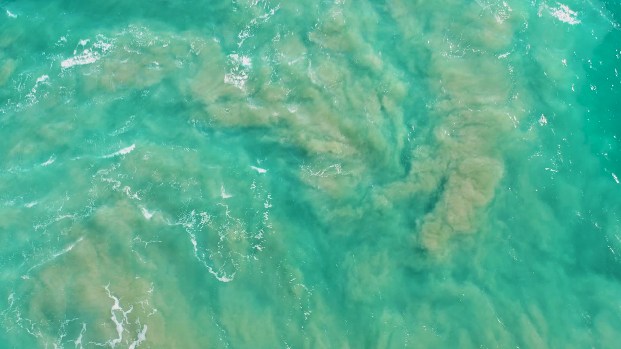 Rippling turquoise water in San Jose del Cabo fills the frame, with tan sandy patches below and brighter foam marks toward the left, no visible people.